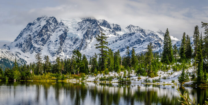 A lake reflects the image of a snow covered mountain and moraine lined with evergreen trees.